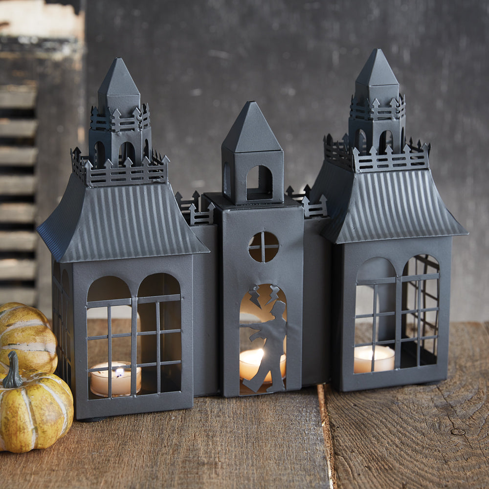 Three gray house-shaped lanterns with candles inside on a wooden surface.