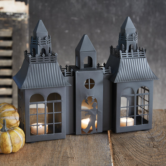 Three gray house-shaped lanterns with candles inside on a wooden surface.